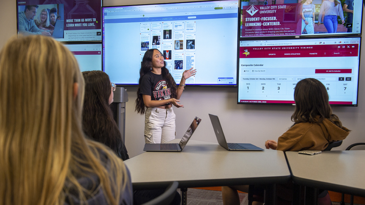 Female student giving a speaking to a class with tv screens behind