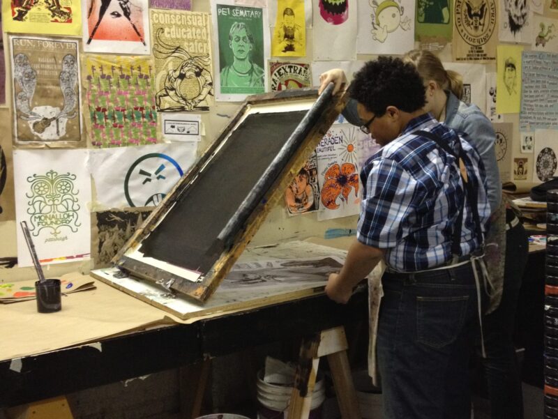 Two artists work on creating a print at a table in front of a wall filled with prints made by other artists.