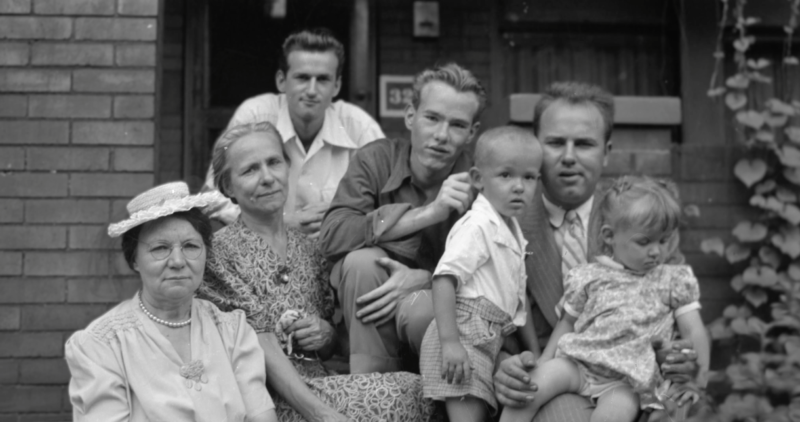 Black and white film still. A teenage Andy Warhol sits on a stoop, surrounded by family members.