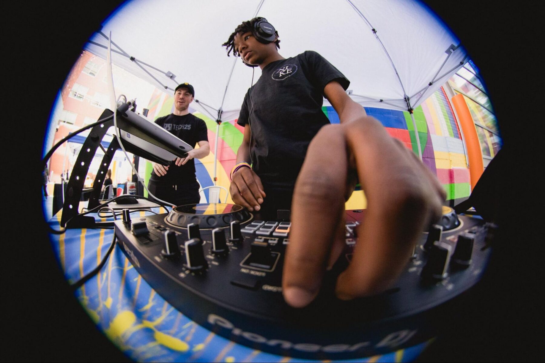 a DJ adjusting knobs and sliders on a Pioneer DJ mixer at an outdoor music event. Another person is partially visible in the background under a tent with colorful patterns. The perspective is from behind the DJ booth.