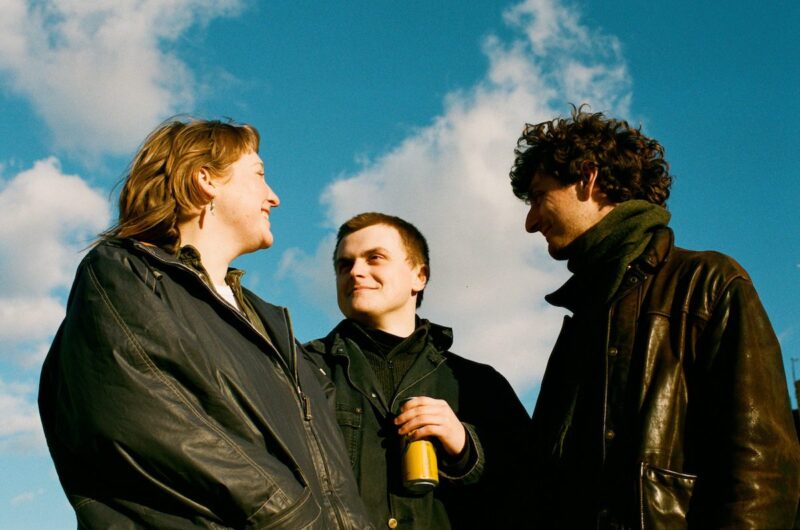 Three people in black coats standing outdoors and smiling under a clear blue sky.