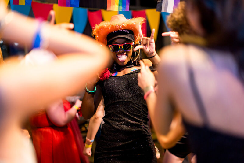 Person wearing an orange hat and sunglasses dances joyfully at a lively party with colorful decorations in the background.