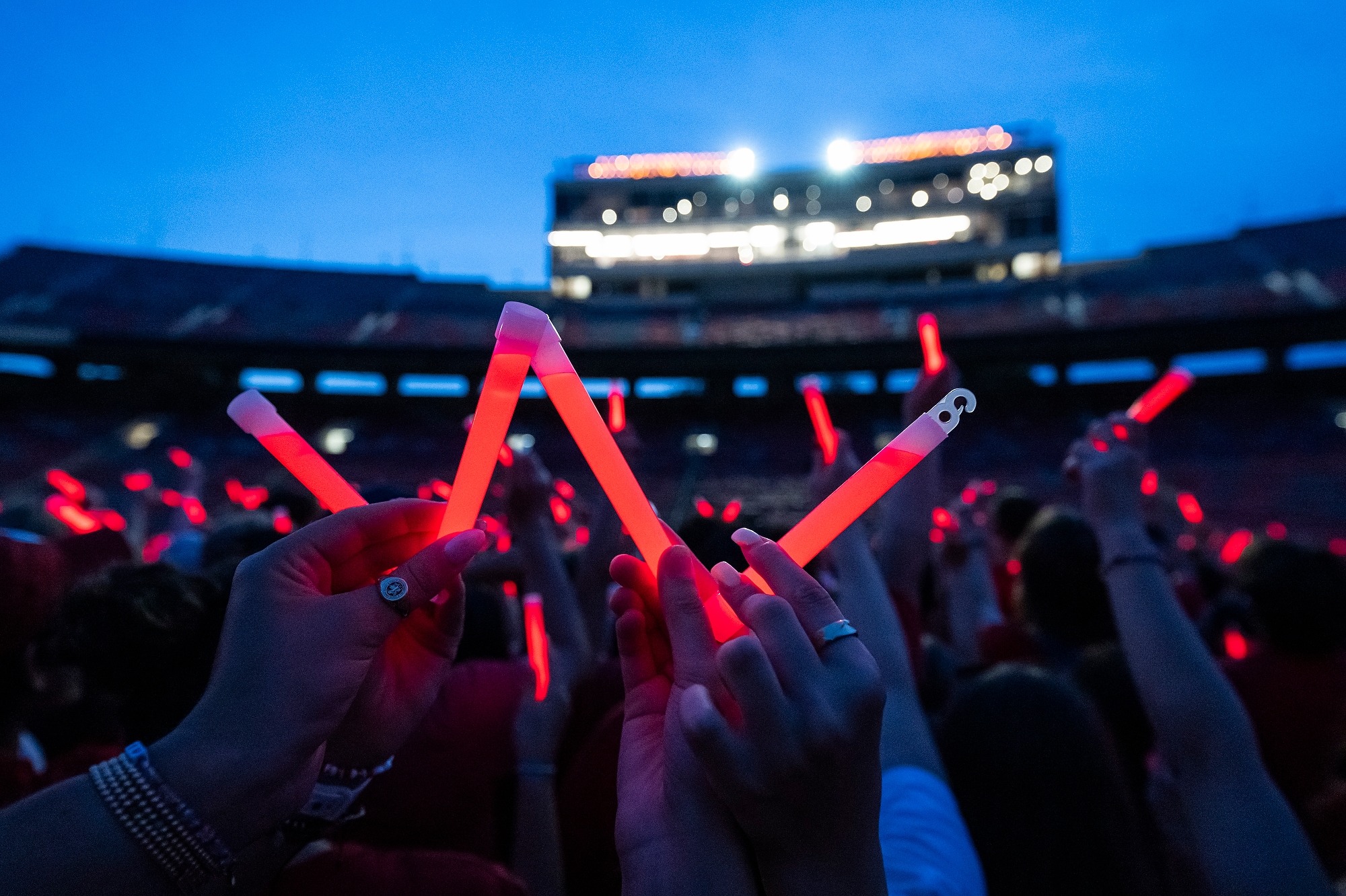 Audience members holding up glowing red sticks at a nighttime event in Camp Randall Stadium. In the foreground, someone holds the sticks to form a letter W.