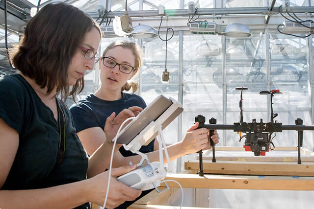 Two students are working with a drone and tablet computer inside a greenhouse