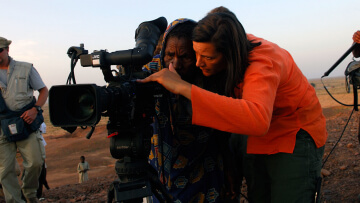 A woman filmmaker in an orange shirt shows another woman in ethnic garb the film camera, emphasizing WMM's mission of empowering women filmmakers all over the world